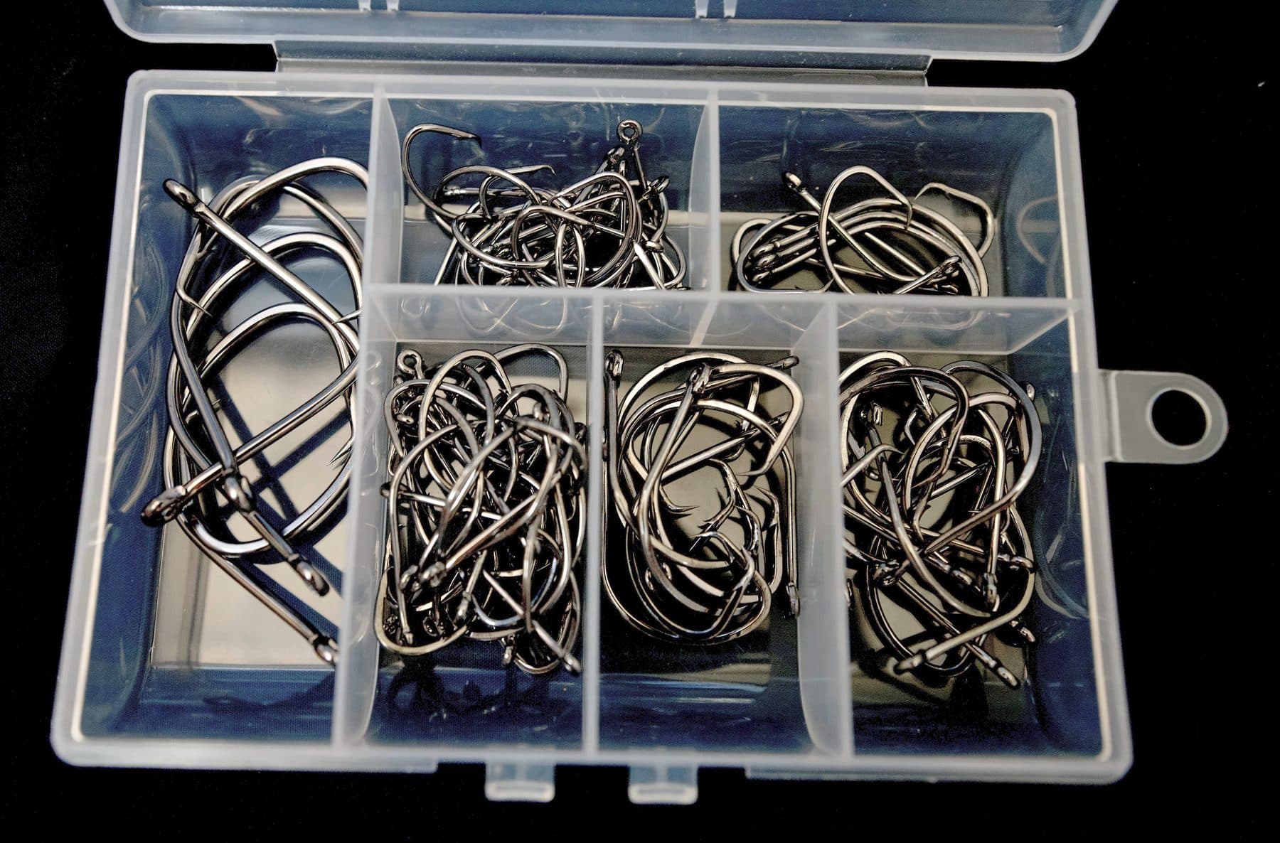 Open clear six-compartment tackle box of black nickel circle hooks sorted by size on a black background