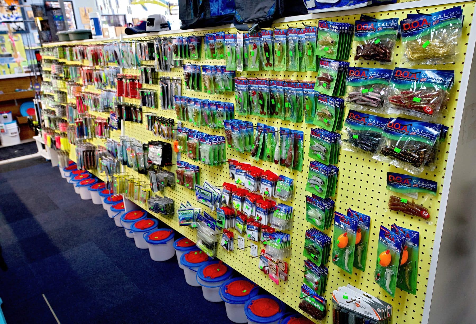 Long yellow pegboard wall of D.O.A. C.A.L. Series soft plastics, Bait Buster lures and Deadly Combo rigs above bait buckets