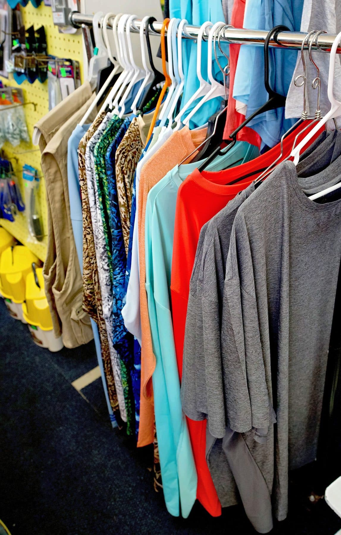Chrome rack of fishing shirts in gray, red, seafoam, orange and camo prints beside a khaki fishing vest