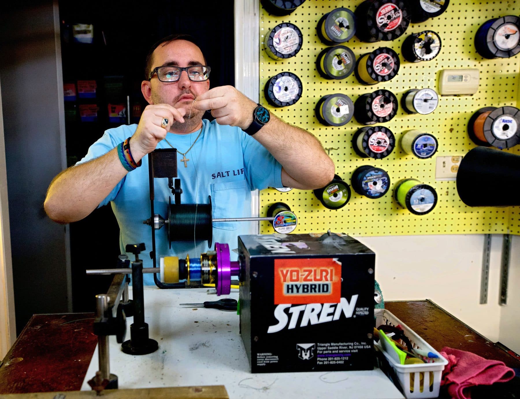 Crew member in glasses threading line at a Stren spooling machine, wall of bulk braid and mono spools behind