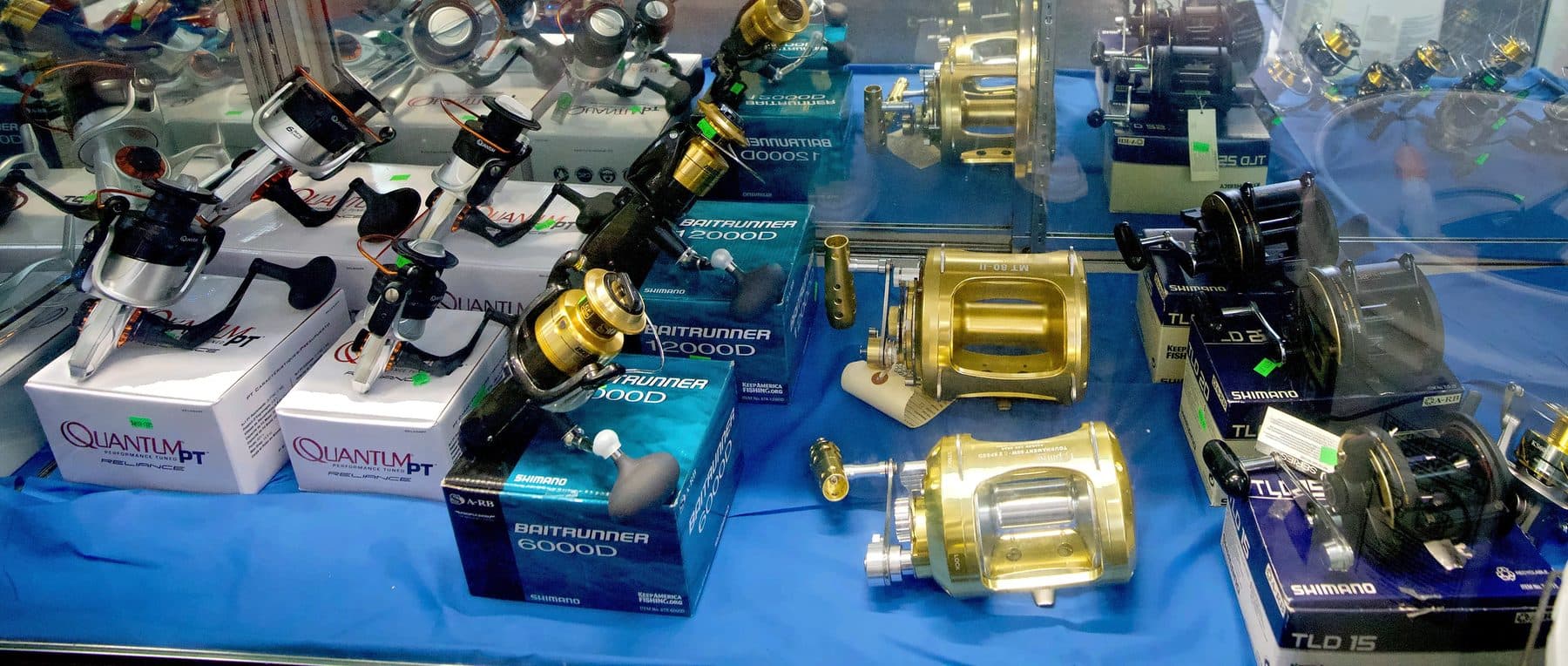 Glass case of boxed Shimano Baitrunner and TLD reels, Quantum PT reels and gold two-speed offshore reels on blue cloth